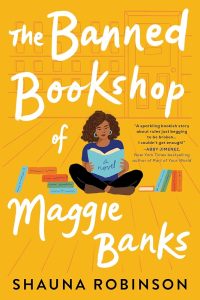 The cover of the novel The Banned Bookshop of Maggie Banks by Shauna Robinson. The cover is yellow and features a young Black woman sitting on the floor reading a book. There are stacks of books on either side of her. 